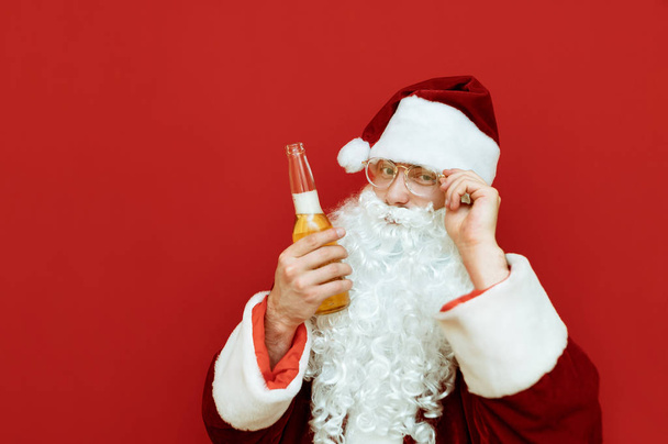 Nahaufnahme Porträt eines fröhlichen Weihnachtsmannes mit einer Bierflasche in der Hand, justiert seine Brille, blickt in die Kamera und lächelt. Lustiger Weihnachtsmann trinkt Alkohol und posiert vor der Kamera. - Foto, Bild