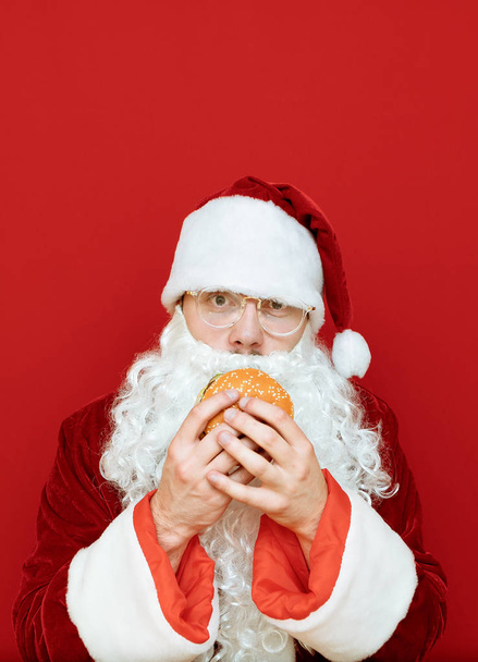 Kırmızı kostümlü ve kırmızı arka planda sakallı Noel Baba elinde lezzetli bir burger tutuyor ve yemek üzere. Komik Noel Baba arka planda fast food yiyor. Dikey fotoğraf. - Fotoğraf, Görsel