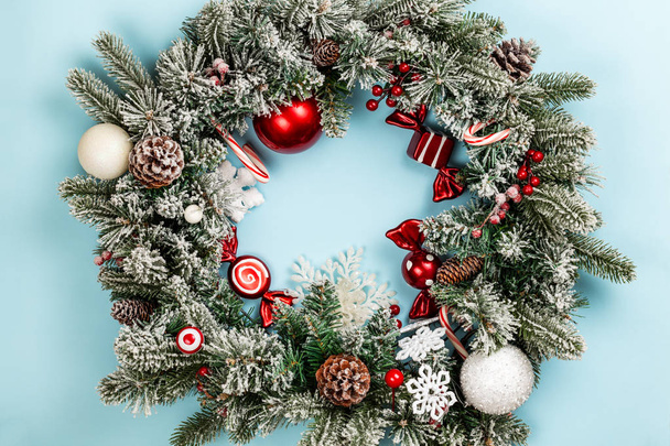 Christmas wreath or crown composition with frozen fir garlands, spheres, sweets and candy cane over a blue background. Flat lay, top view with copy space. - Φωτογραφία, εικόνα