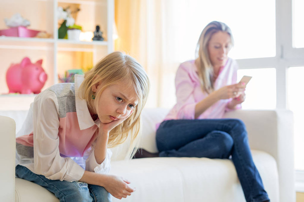Mother is texting while her little girl  unhappy sitting next to her on couch - Foto, imagen