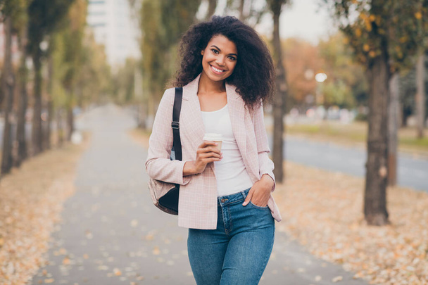 Photo of pretty charming dark skin curly lady walking park after college lectures drink takeaway hot coffee hold backpack warm fall season wear jacket jeans outdoors - Foto, Imagem