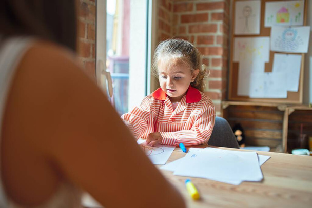 Beautiful teacher and blond student toddler girl wearing school uniform drawing draw using colored pens at kindergarten - Photo, image