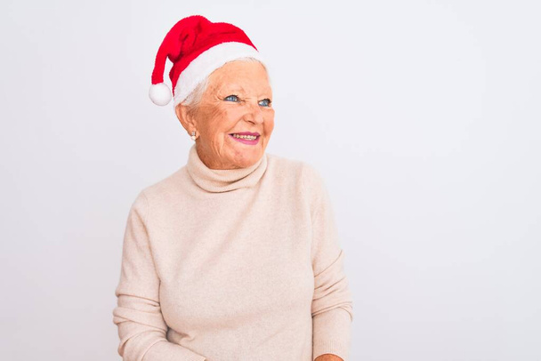 Idős ősz hajú nő, Crhistmas Mikulás kalapot visel elszigetelt fehér háttér felett, oldalra néz mosollyal az arcán, természetes kifejezéssel. Nevetve magabiztos. - Fotó, kép
