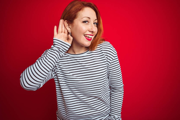 Jonge roodharige vrouw draagt bandjes marineblauw shirt dat over rood geïsoleerde achtergrond staat te glimlachen met hand op oor luisterend naar geruchten of roddels. Doofheid. - Foto, afbeelding