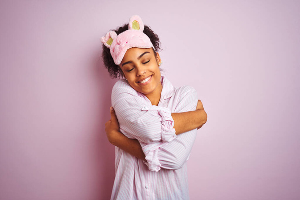 Молодая африканская американка в пижаме и маске на изолированном розовом фоне обнимает себя счастливой и позитивной, уверенно улыбаясь. Самолюбие и забота о себе
 - Фото, изображение