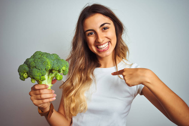 Young beautiful woman eating broccoli over grey isolated background with surprise face pointing finger to himself - Foto, immagini