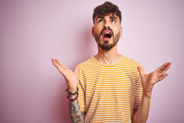 Young man with tattoo wearing yellow striped t-shirt standing over isolated pink background crazy and mad shouting and yelling with aggressive expression and arms raised. Frustration concept. - Photo, Image