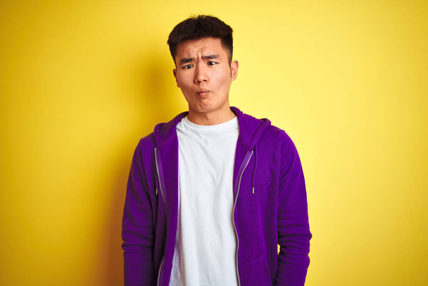 Joven asiático chino hombre vistiendo sudadera púrpura de pie sobre aislado fondo amarillo haciendo cara de pez con labios, loco y cómico gesto. Expresión divertida
. - Foto, imagen