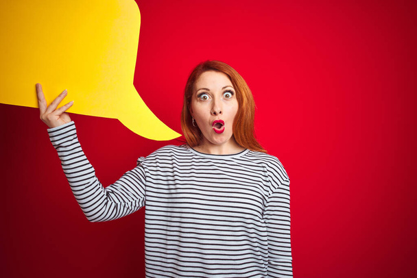 Young beautiful redhead woman holding yellow speech bubble over red isolated background scared in shock with a surprise face, afraid and excited with fear expression - Photo, Image