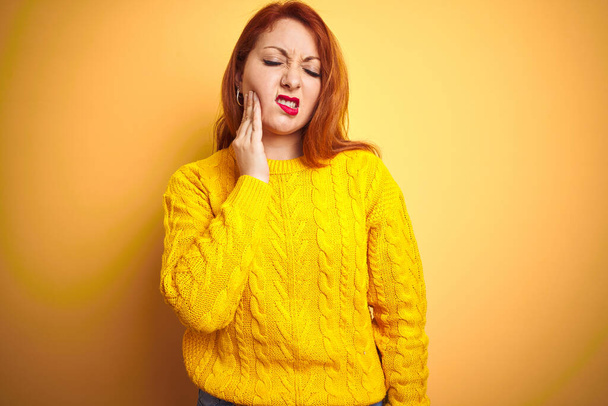 Beautiful redhead woman wearing winter sweater standing over isolated yellow background touching mouth with hand with painful expression because of toothache or dental illness on teeth. Dentist concept. - 写真・画像