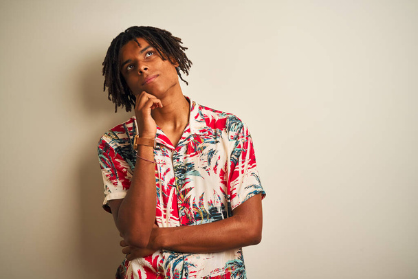 Afro man with dreadlocks on vacation wearing summer shirt over isolated white background with hand on chin thinking about question, pensive expression. Smiling with thoughtful face. Doubt concept. - Fotografie, Obrázek