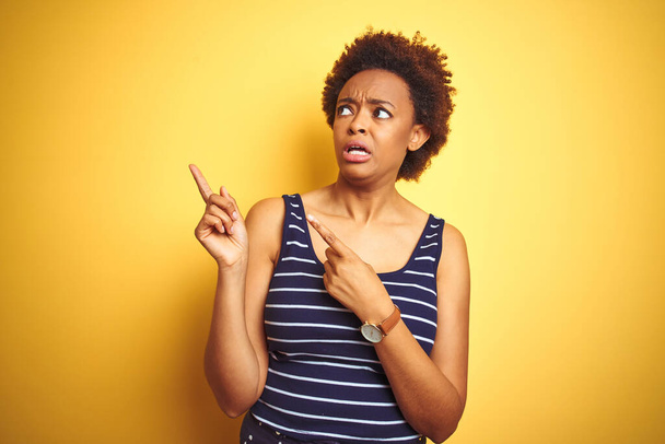 Beauitul african american woman wearing summer t-shirt over isolated yellow background Pointing aside worried and nervous with both hands, concerned and surprised expression - Foto, Imagem