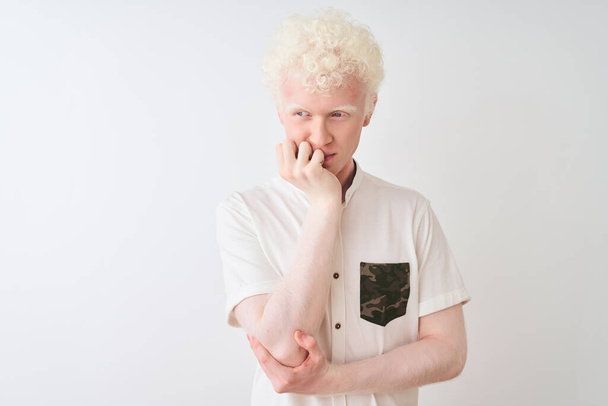 Молодой белокурый альбинос в повседневной рубашке, стоящий на изолированном белом фоне, выглядящий напряженным и нервным с руками во рту, грызущими ногти. Проблема тревоги
. - Фото, изображение
