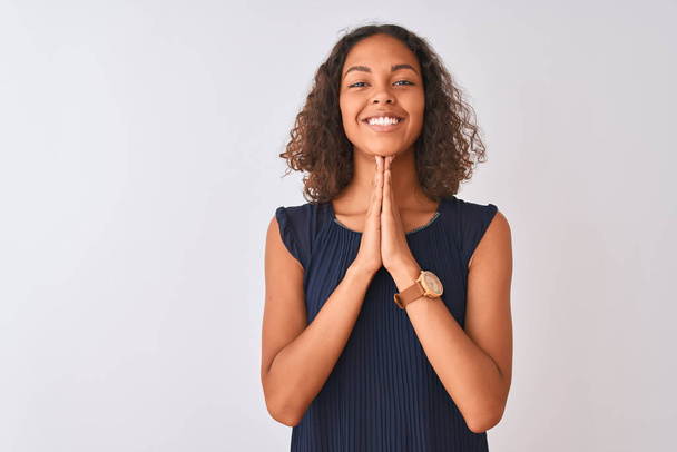Fiatal brazil nő, kék ruhát visel, elszigetelt fehér háttér felett áll, kéz a kézben imádkozik, bocsánatot kér, magabiztosan mosolyog.. - Fotó, kép