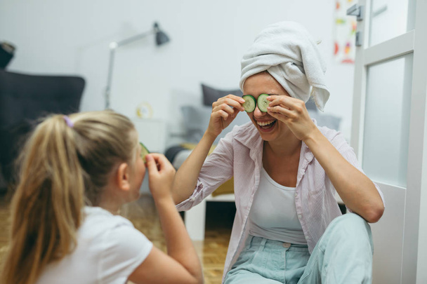 Mother and daughter putting cucumbers on faces at home - Фото, изображение