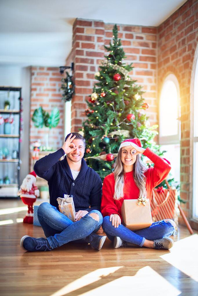 Casal jovem vestindo chapéu de Papai Noel sentado no chão em torno da árvore de natal em casa fazendo gesto ok com a mão sorrindo, olho olhando através dos dedos com o rosto feliz
. - Foto, Imagem