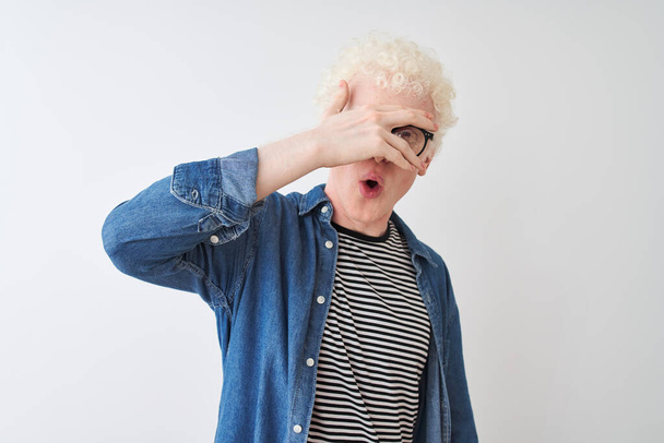 Young albino blond man wearing denim shirt and glasses over isolated white background peeking in shock covering face and eyes with hand, looking through fingers with embarrassed expression. - 写真・画像