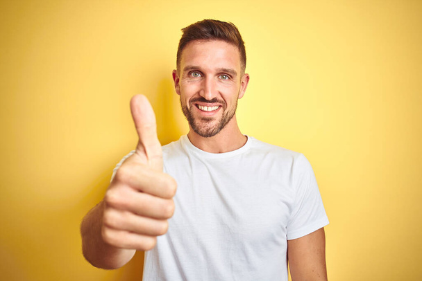 Ein junger gutaussehender Mann trägt ein legeres weißes T-Shirt über gelbem Hintergrund und macht eine glückliche Daumen-hoch-Geste mit der Hand. Zustimmender Blick in die Kamera zeigt Erfolg. - Foto, Bild