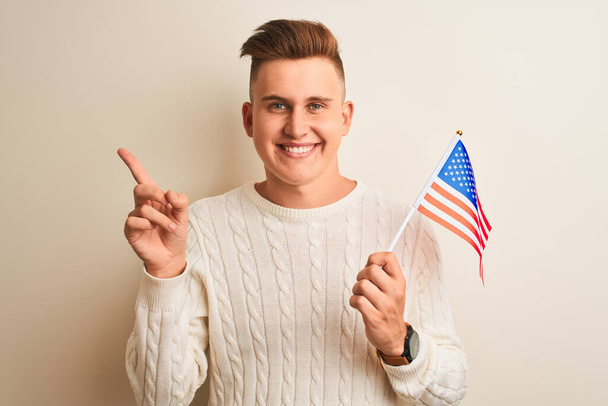 Young handsome man holding USA flag over isolated white background very happy pointing with hand and finger to the side - Foto, Imagen