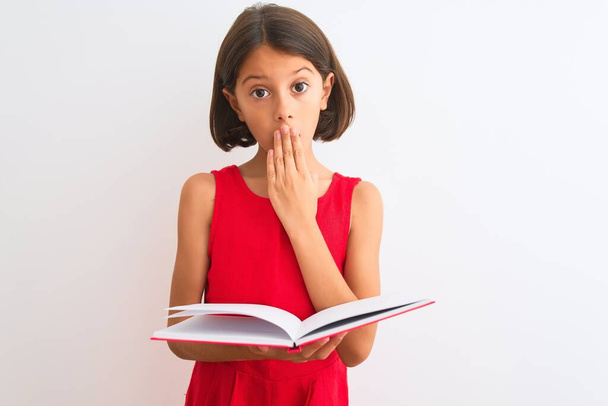 Beautiful student child girl reading red book standing over isolated white background cover mouth with hand shocked with shame for mistake, expression of fear, scared in silence, secret concept - Фото, зображення