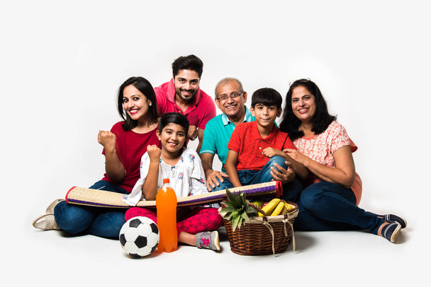 インドの家族は屋内でピクニックを楽しんでいます-フルーツバスケット、マット、サッカー、飲み物で白い背景に座っているアジアの家族の多世代。選択的焦点 - 写真・画像