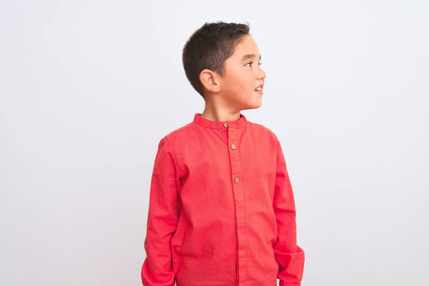 schöner kleiner Junge in elegantem rotem Hemd, der vor isoliertem weißem Hintergrund steht und zur Seite schaut, mit einem Lächeln im Gesicht, natürlichem Ausdruck. Lachen selbstbewusst. - Foto, Bild