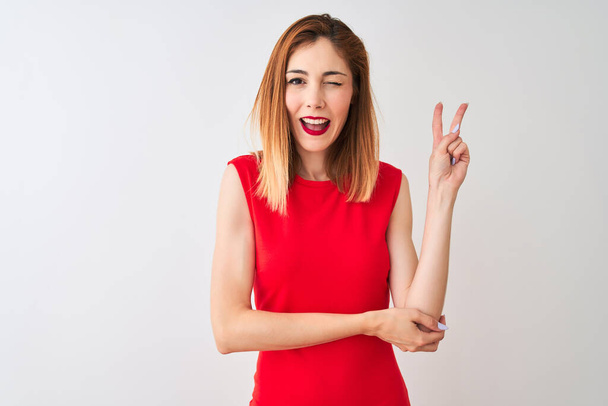 Femme d'affaires rousse portant une élégante robe rouge debout sur un fond blanc isolé souriant avec un visage heureux clin d'oeil à la caméra faisant signe de victoire avec les doigts. Numéro deux
. - Photo, image