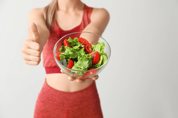 Sporty woman with healthy vegetable salad showing thumb-up gesture on light background, closeup. Diet concept - Photo, Image