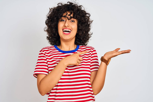 junge arabische Frau mit lockigem Haar trägt gestreiftes T-Shirt über isoliertem weißem Hintergrund erstaunt und lächelt in die Kamera, während sie mit der Hand präsentiert und mit dem Finger zeigt. - Foto, Bild