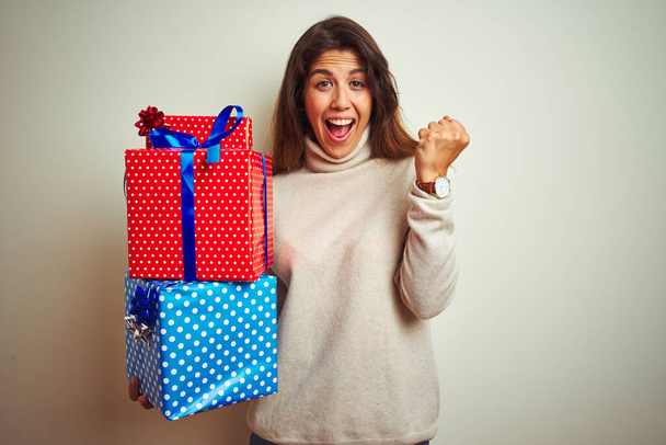 Young beautiful woman holding birthday gifts standing over isolated white background screaming proud and celebrating victory and success very excited, cheering emotion - 写真・画像