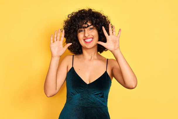 Jeune femme arabe aux cheveux bouclés portant une robe élégante sur fond jaune isolé montrant et pointant vers le haut avec les doigts numéro dix tout en souriant confiant et heureux
. - Photo, image