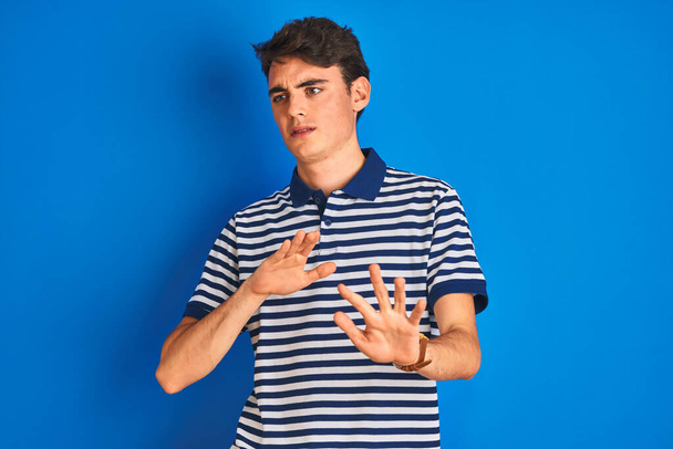 Teenager αγόρι φορώντας casual t-shirt στέκεται πάνω από το μπλε απομονωμένο φόντο αηδιασμένη έκφραση, δυσαρεστημένος και φοβισμένος κάνει αηδία πρόσωπο επειδή αντίδραση αποστροφή. Με τα χέρια ψηλά. Ενοχλητική έννοια. - Φωτογραφία, εικόνα