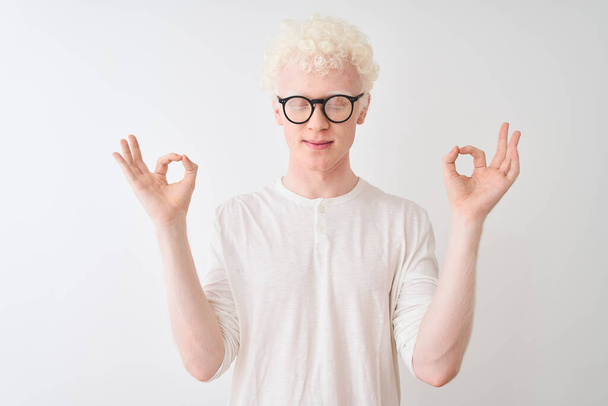 junger albinoblonder Mann mit T-Shirt und Brille, der vor isoliertem weißem Hintergrund steht, entspannt und lächelnd mit geschlossenen Augen Meditationsgeste mit den Fingern macht. Yoga-Konzept. - Foto, Bild