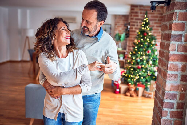 Красивая пара средних лет улыбается счастливой и уверенной. Стоять и танцевать вокруг новогодней елки дома
 - Фото, изображение