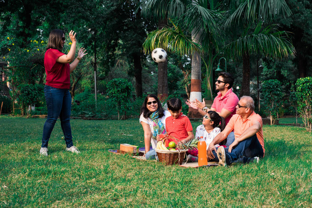 Indian Family enjoying Picnic - Multi generation of asian family sitting over lawn or green grass in park with fruit basket, mat and drinks. selective focus - Photo, Image