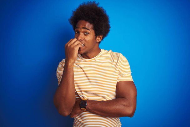 amerikanischer Mann mit Afro-Haaren, der ein gestreiftes gelbes T-Shirt über isoliertem blauem Hintergrund trägt und gestresst und nervös aussieht, die Hände auf mundgebissenen Nägeln. Angstproblem. - Foto, Bild