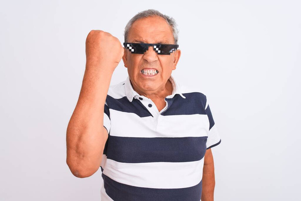 Senior grijsharige man draagt gestreepte polo en zonnebril over geïsoleerde witte achtergrond geïrriteerd en gefrustreerd schreeuwen van woede, gek en schreeuwen met opgeheven hand, woede concept - Foto, afbeelding