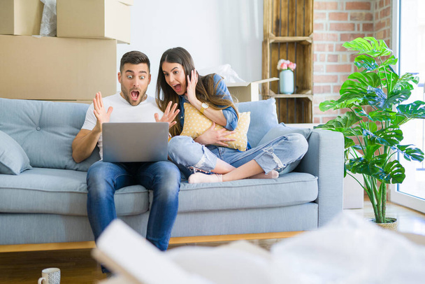 junges schönes Paar entspannt auf dem Sofa des neuen Hauses mit Computer-Laptop sehr glücklich und aufgeregt, Siegerausdruck feiert Sieg schreiend mit breitem Lächeln und erhobenen Händen - Foto, Bild