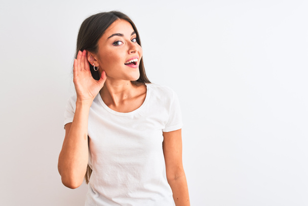 junge schöne Frau in lässigem T-Shirt, die vor isoliertem weißem Hintergrund steht und lächelnd mit der Hand über dem Ohr Gerüchten oder Klatsch lauscht. Konzept für Gehörlosigkeit. - Foto, Bild