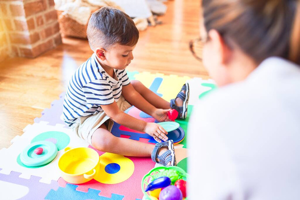 Красивий маленький хлопчик сидить на головоломці, граючи з пластиковими тарілками, фруктами та овочами в дитячому саду
 - Фото, зображення