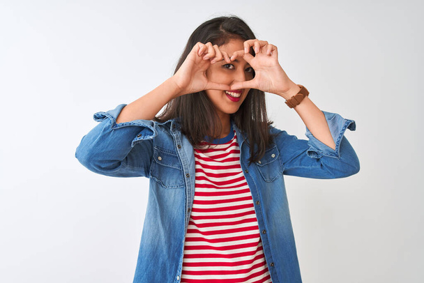 Молода жінка в смугастій футболці і денім сорочці над ізольованим білим фоном Виконання форми серця з рукою і пальцями посміхається дивлячись через знак - Фото, зображення