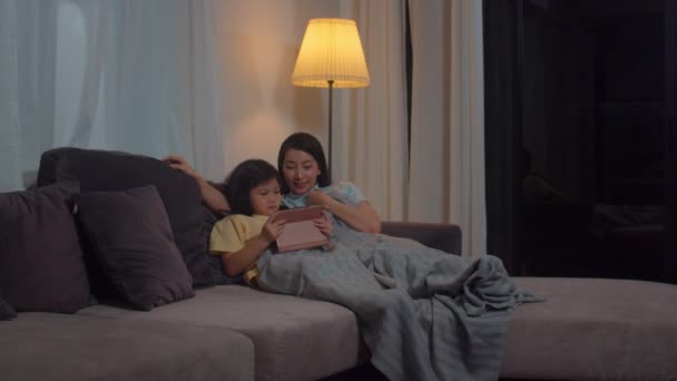 Joven familia asiática y su hija feliz usando la tableta en casa. madre coreana relajarse con la niña viendo la película tumbada en el sofá antes de ir a dormir en la sala de estar en casa por la noche
. - Metraje, vídeo