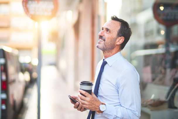 Middelbare leeftijd knappe zakenman met behulp van smartphone drinken take away koffie glimlachen - Foto, afbeelding
