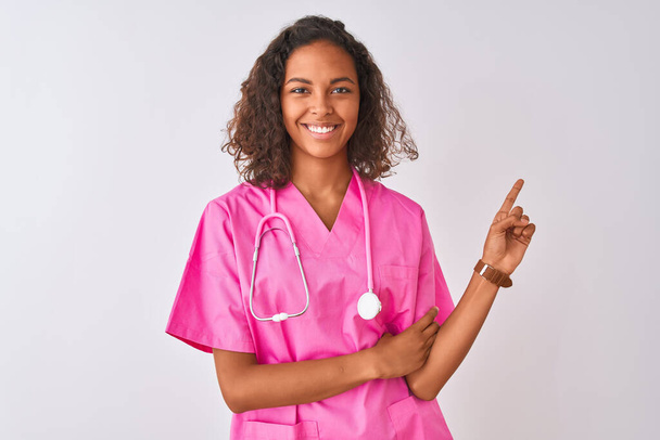Młoda brazylijska pielęgniarka nosząca stetoskop stojąca nad odizolowanym białym tłem z dużym uśmiechem na twarzy, wskazująca ręką i palcem na bok patrzącą w kamerę. - Zdjęcie, obraz