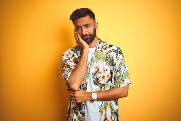 junger indischer Mann im Urlaub mit Sommerblumenhemd vor isoliertem gelben Hintergrund, der müde und gelangweilt mit Depressionsproblemen mit verschränkten Armen aussieht. - Foto, Bild