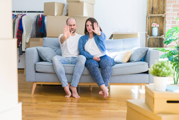 Young couple sitting on the sofa arround cardboard boxes moving to a new house doing stop sing with palm of the hand. Warning expression with negative and serious gesture on the face. - Photo, Image