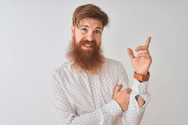 junger rothaariger irischer Mann im Hemd vor isoliertem weißem Hintergrund mit einem breiten Lächeln im Gesicht, der mit der Hand und dem Finger zur Seite in die Kamera zeigt. - Foto, Bild
