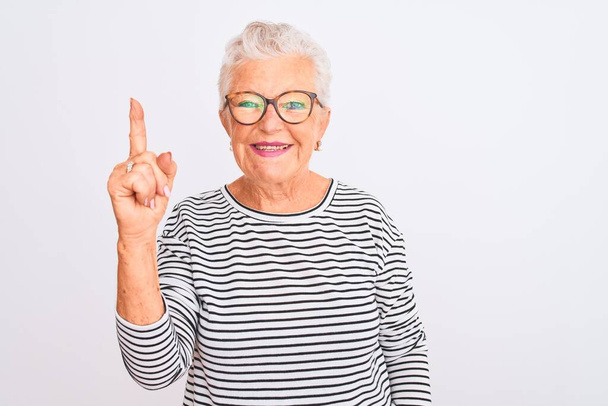 Senior harmaa tukka nainen yllään raidallinen laivaston t-paita lasit yli eristetty valkoinen tausta näyttää ja osoittaa sormella numero yksi hymyillen itsevarma ja onnellinen
. - Valokuva, kuva