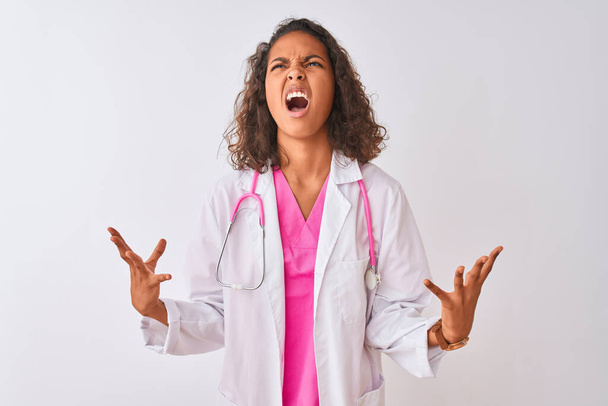 Молодая бразильская докторша в стетоскопе, стоящая на изолированном белом фоне, сумасшедшая, кричащая и кричащая с агрессивным выражением лица и поднятыми руками. Концепция разочарования
. - Фото, изображение