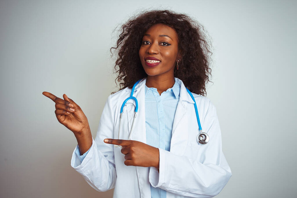 Fiatal afro-amerikai orvos nő visel sztetoszkóppal izolált fehér háttér mosolyog, és néz a kamera mutat két kézzel és ujjal oldalra. - Fotó, kép